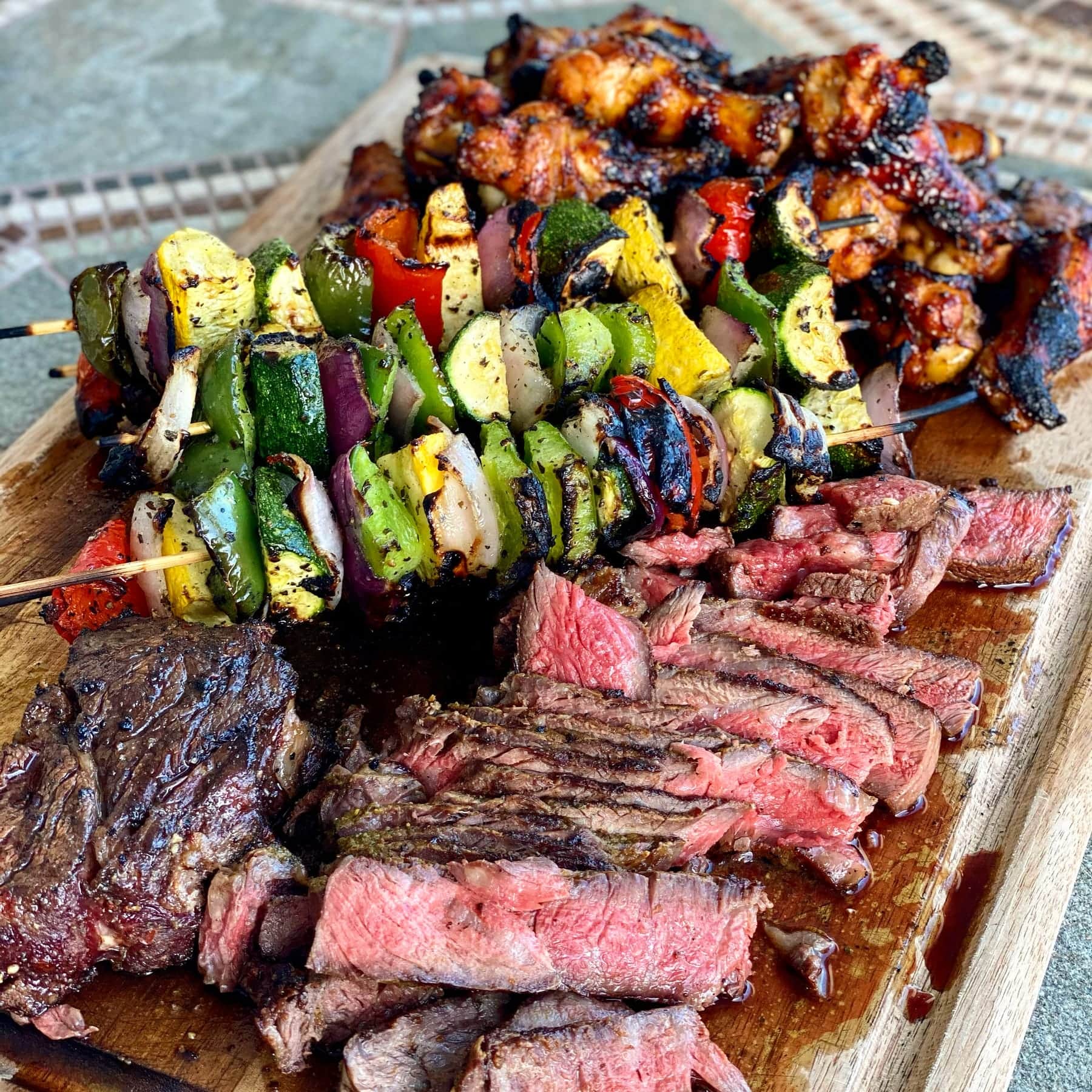 Grilled vegetable skewers over sliced steak on a wooden board